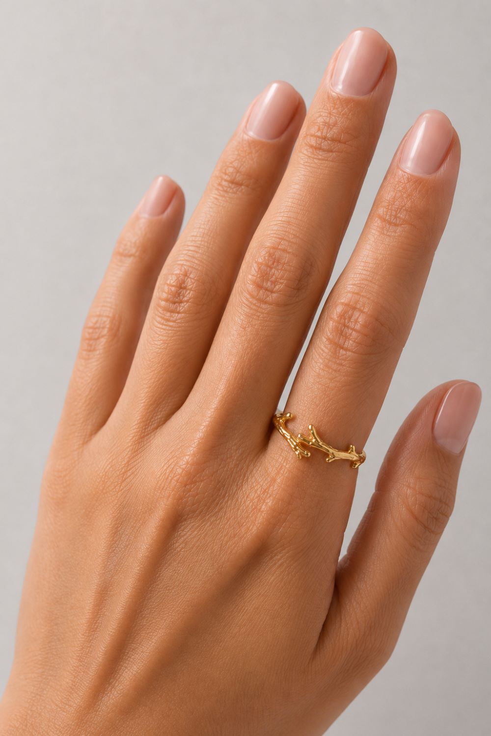 A close-up of a hand wearing a delicate gold ring with a small branch or leaf motif, set against a plain light gray background.