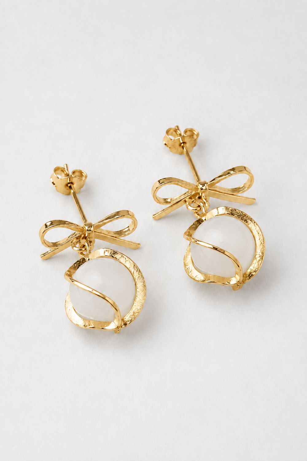 A pair of gold-toned dangle earrings featuring a textured bow and a twisted oval drop, displayed on a clean light gray background.