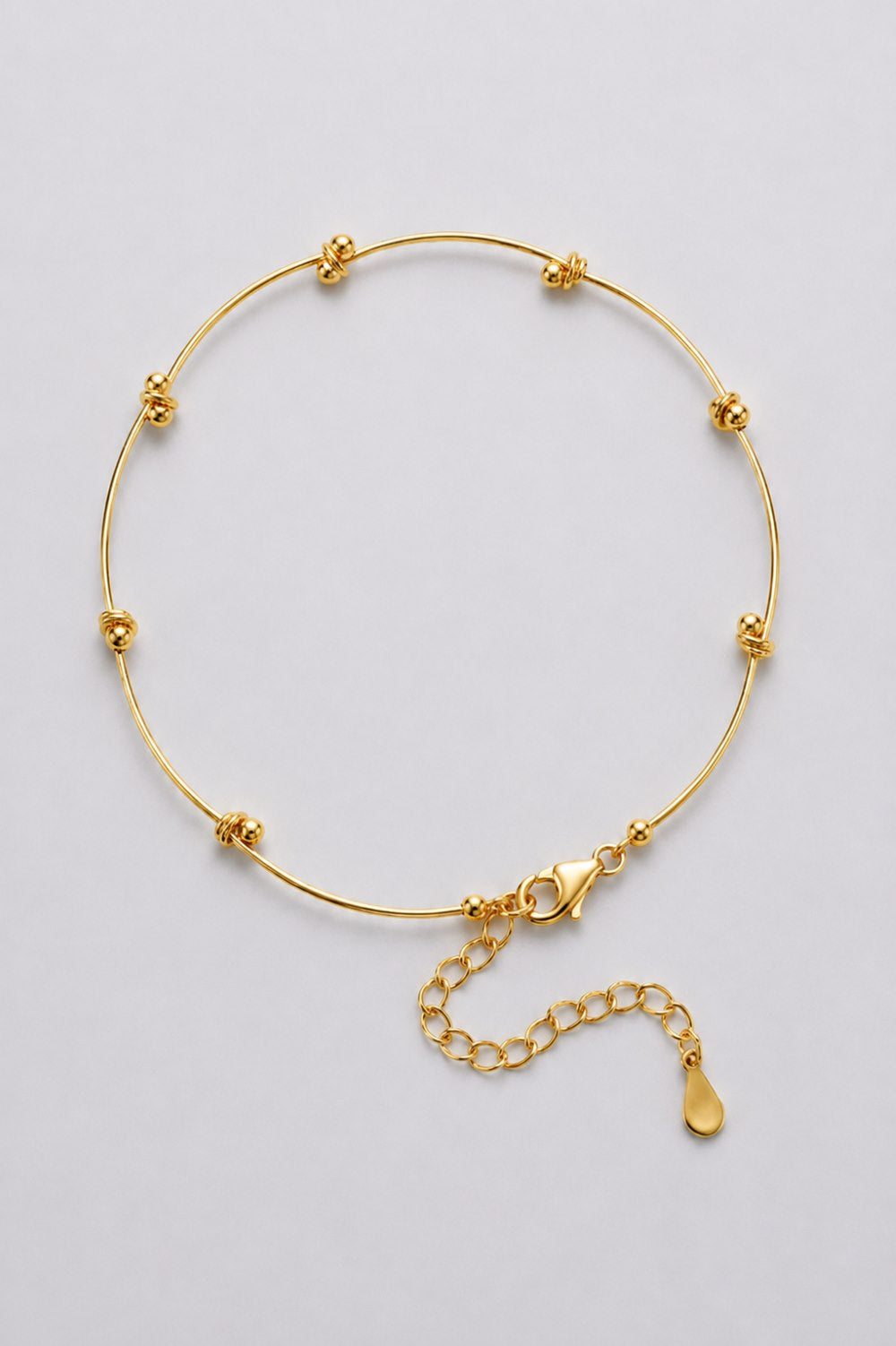 A delicate gold bangle bracelet with small flower-shaped beads spaced along the thin band, a lobster clasp, and an adjustable chain extender with a teardrop charm, shown on a light gray background.