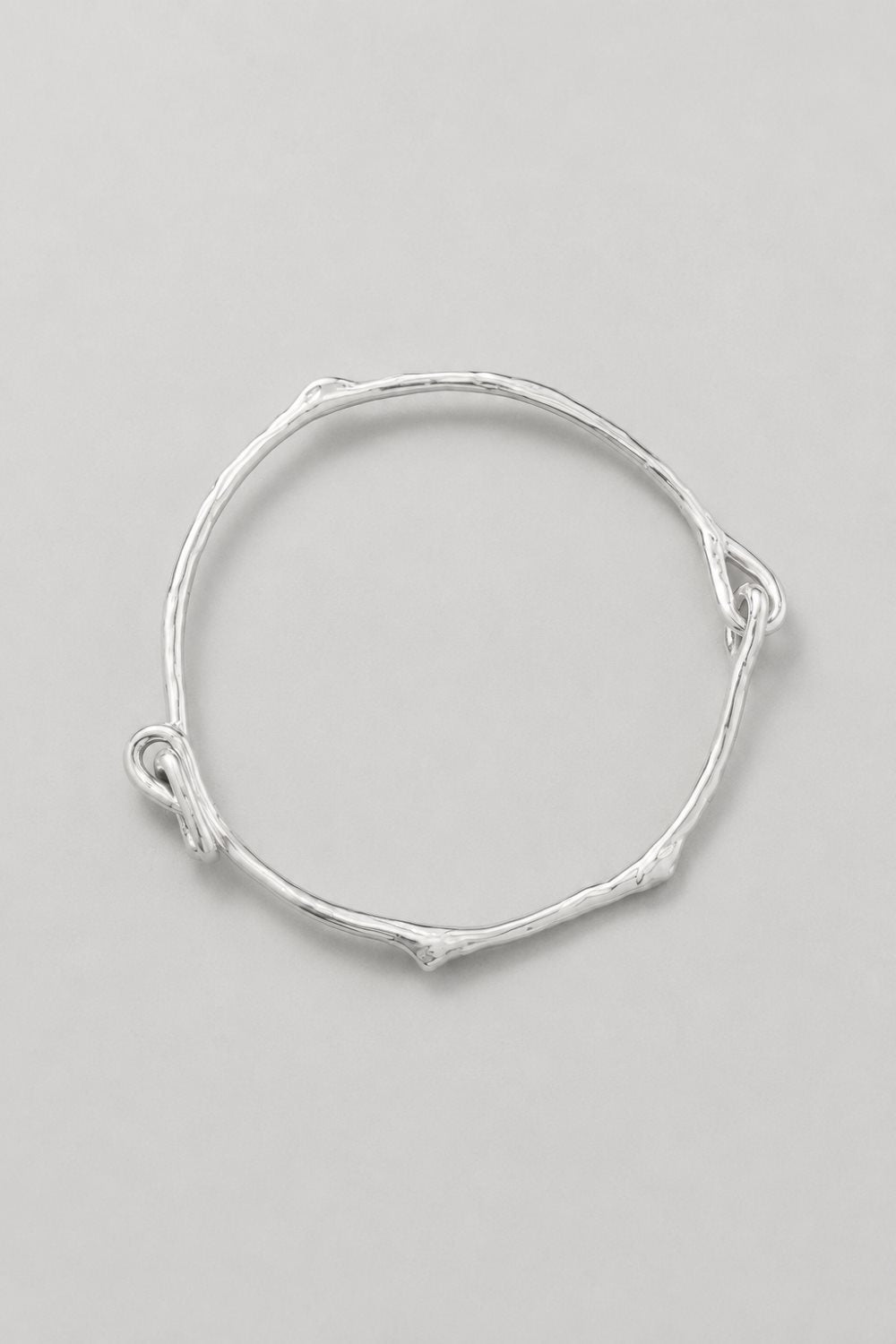 A thin silver bangle bracelet with a textured, twig-like organic shape and small loop details at the ends, displayed on a neutral light gray background.
