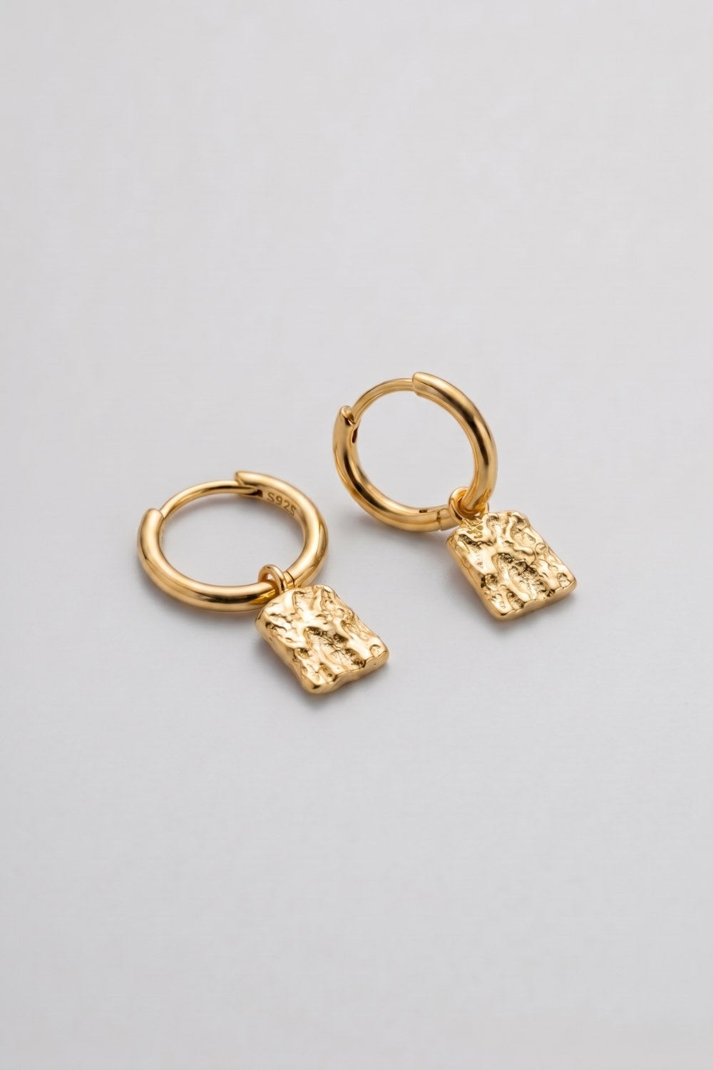 A pair of gold huggie hoop earrings with textured, irregularly shaped dangling charms, displayed on a clean light gray background.