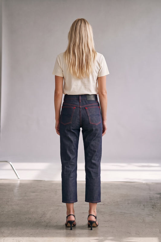 A woman viewed from behind wearing dark indigo cropped jeans with red contrast stitching, a cream t-shirt, and black sandals in a minimalist studio setting.