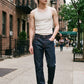 A man walking on a city sidewalk wearing a cream ribbed tank top, dark blue jeans, black boots, and sunglasses.
