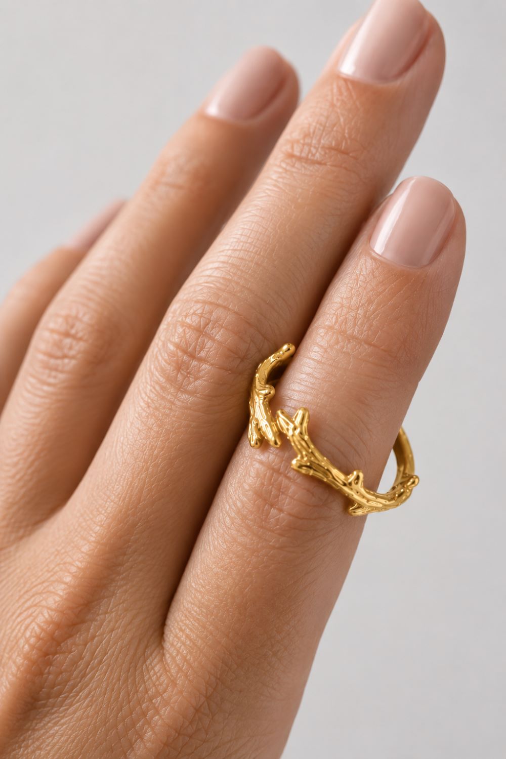 A close-up of a hand wearing a textured gold ring designed to look like a small branch or antler, set against a soft light gray background.
