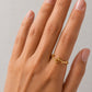 A close-up of a hand wearing a delicate gold ring with a small branch or leaf motif, set against a plain light gray background.