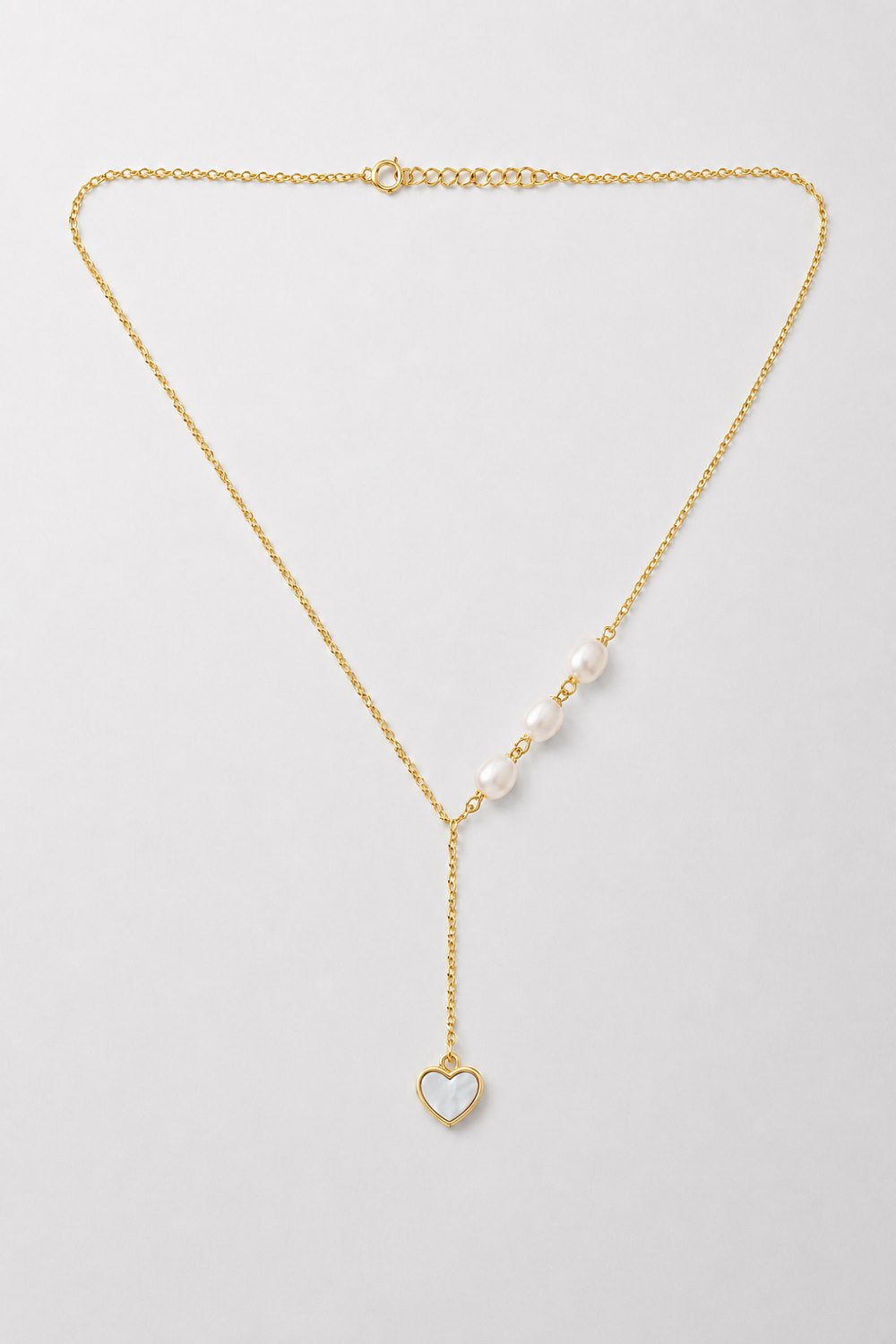A delicate gold Y-necklace featuring a row of white pearls on one side and a small gold heart pendant with a white inlay on the other, displayed on a light gray background.