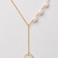A delicate gold Y-necklace featuring a row of white pearls on one side and a dangling gold heart pendant with a white inlay on the other, displayed on a light gray background.