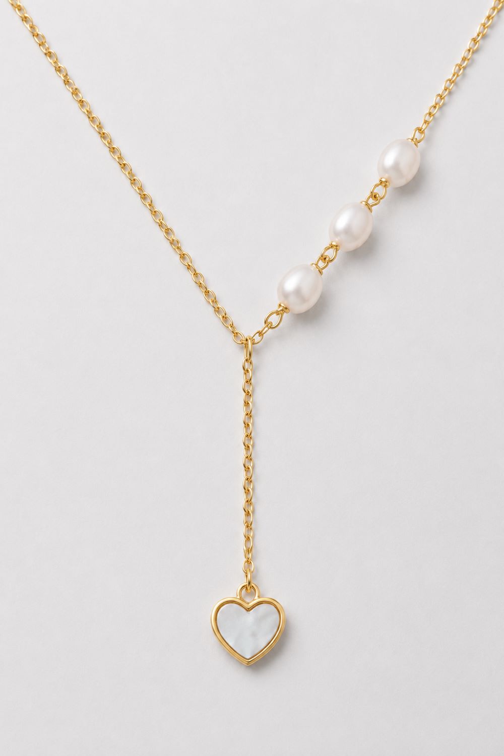 A delicate gold Y-necklace featuring a row of white pearls on one side and a dangling gold heart pendant with a white inlay on the other, displayed on a light gray background.