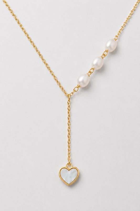 A delicate gold Y-necklace featuring a row of white pearls on one side and a dangling gold heart pendant with a white inlay on the other, displayed on a light gray background.