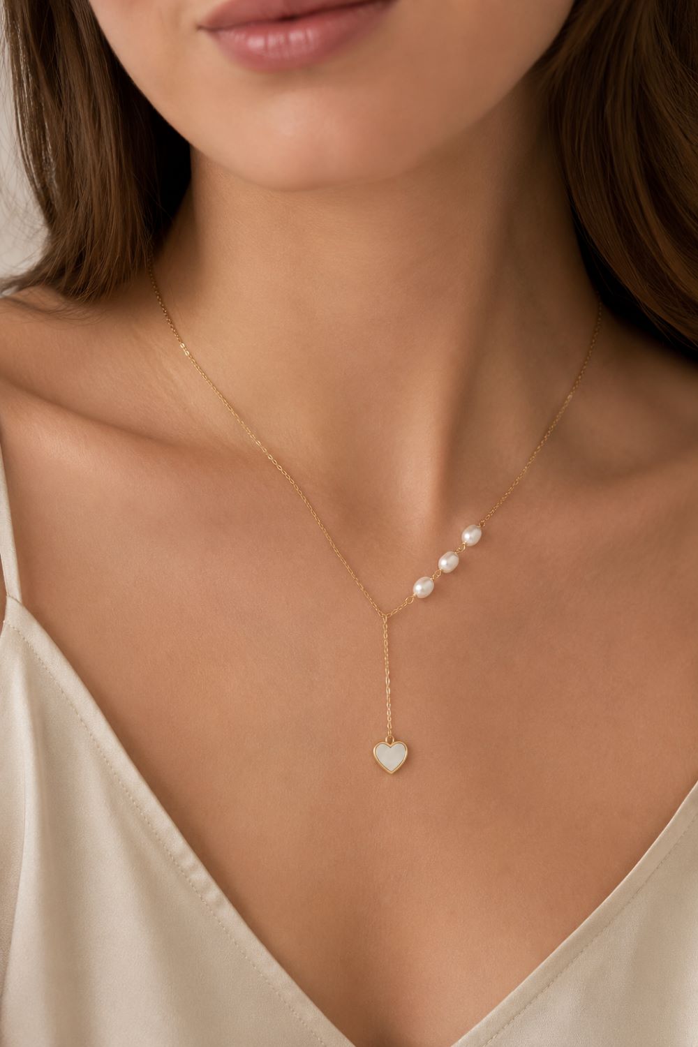 A close-up of a model wearing a delicate gold Y-necklace with three small white pearls and a heart-shaped pearl pendant, paired with a cream satin top.