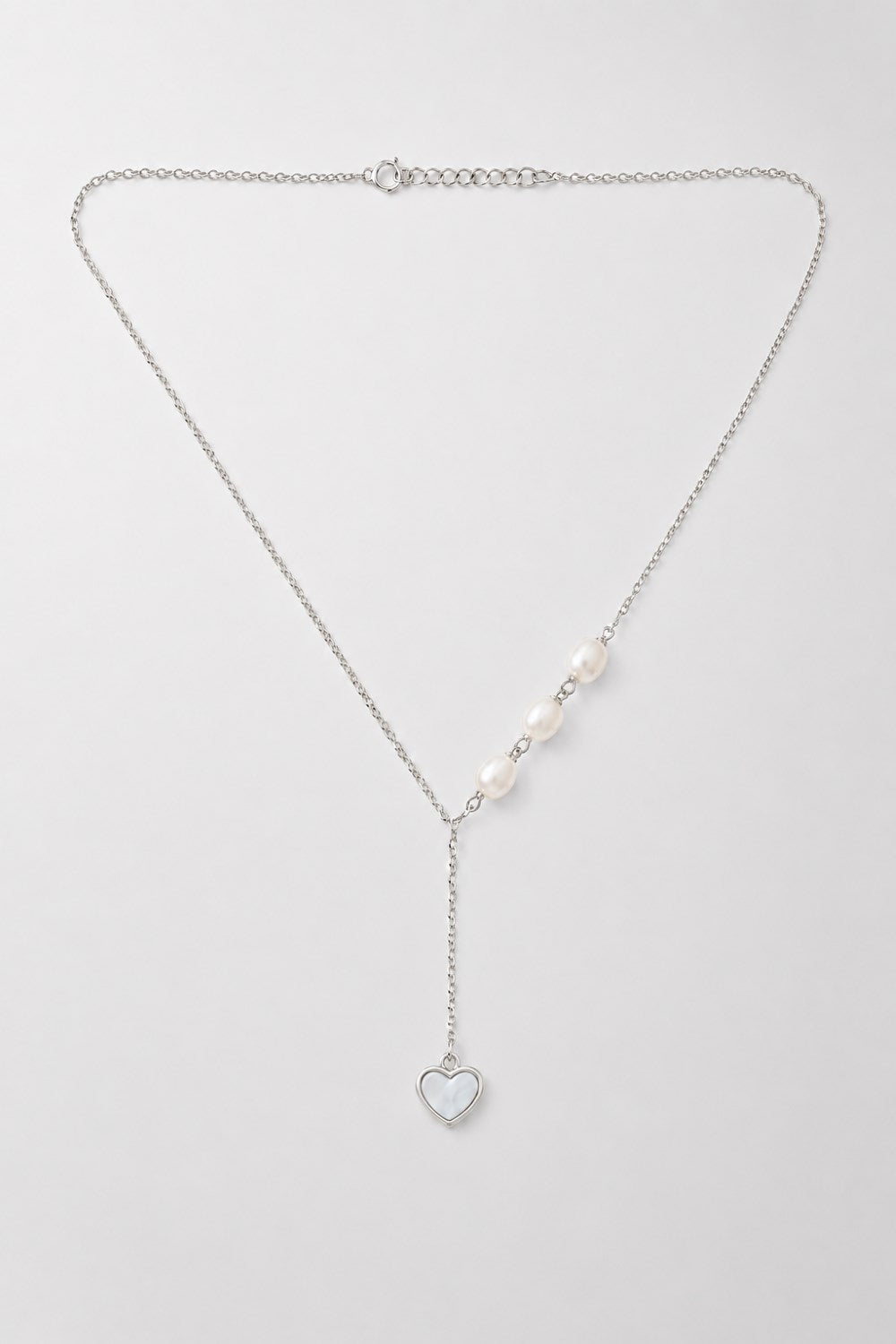 A delicate silver Y-necklace featuring a central row of white pearls and a dangling silver heart charm, displayed on a clean light gray background.