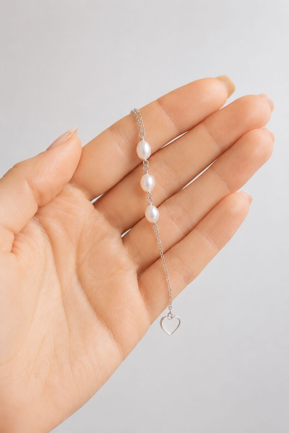 A delicate silver chain bracelet with three white freshwater pearls and a small open heart charm, held in a person's hand against a light gray background.