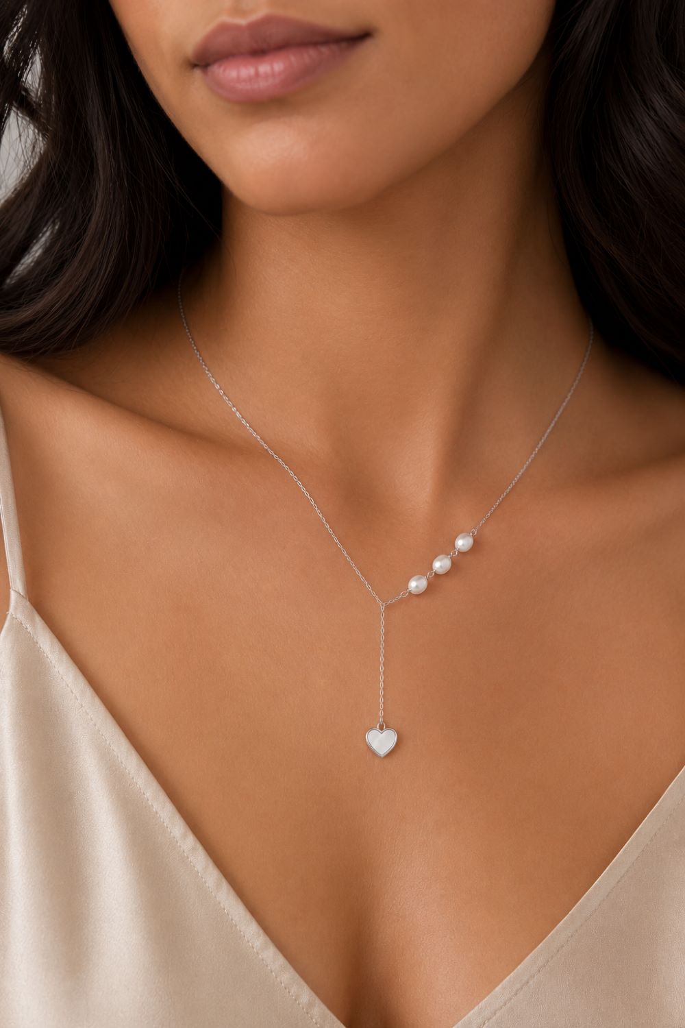 A close-up of a woman wearing a delicate silver Y-necklace with three small pearls and a heart-shaped pendant, paired with a beige satin top.
