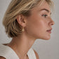 A close-up side profile of a woman with short blonde hair wearing a delicate gold dangle earring with a circular textured charm, set against a neutral gray background.