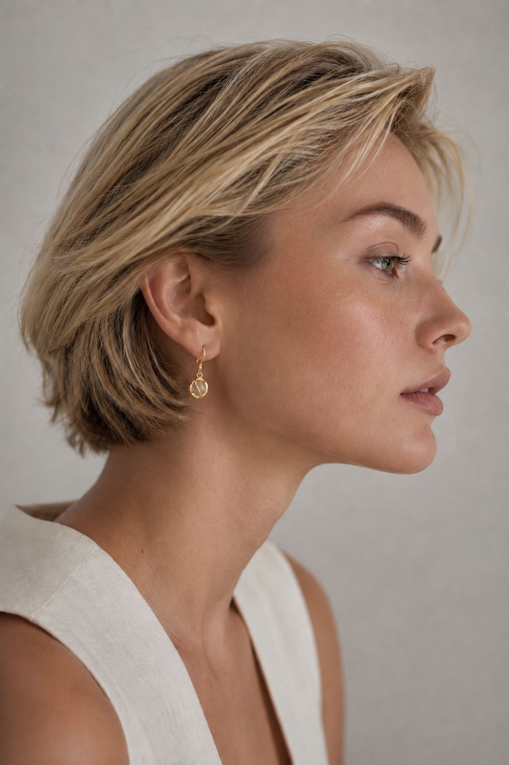 A close-up side profile of a woman with short blonde hair wearing a delicate gold dangle earring with a circular textured charm, set against a neutral gray background.