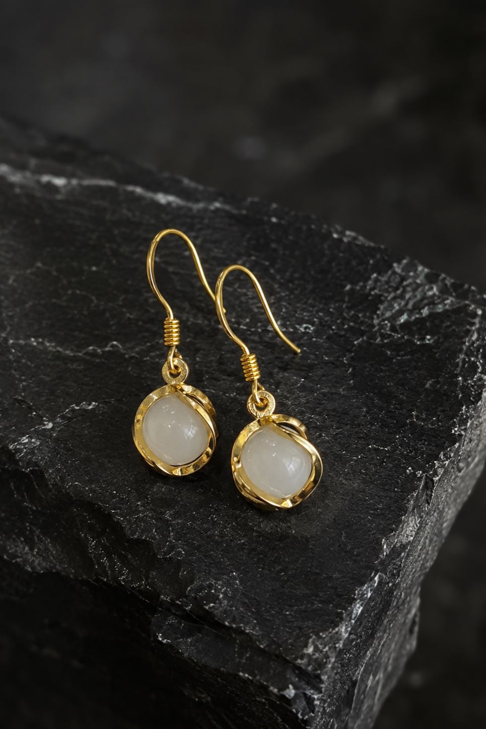 A pair of gold drop earrings with irregular cream-colored stone pendants, displayed on a dark textured slate surface.