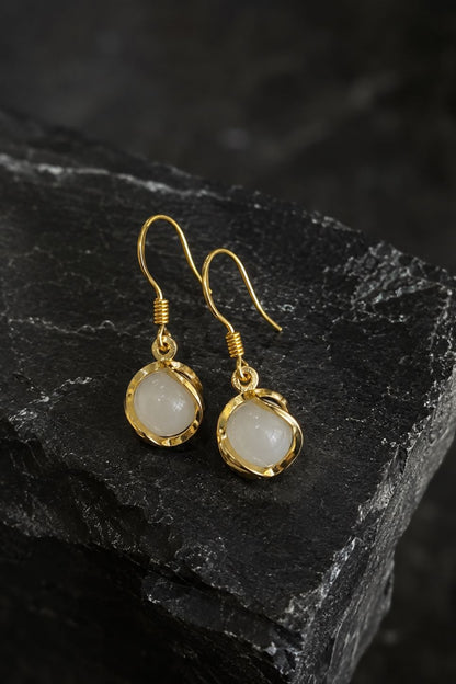 A pair of gold drop earrings with irregular cream-colored stone pendants, displayed on a dark textured slate surface.