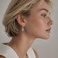 A close-up side profile of a woman with short blonde hair wearing a delicate silver drop earring with a clear gemstone, set against a neutral light gray background.