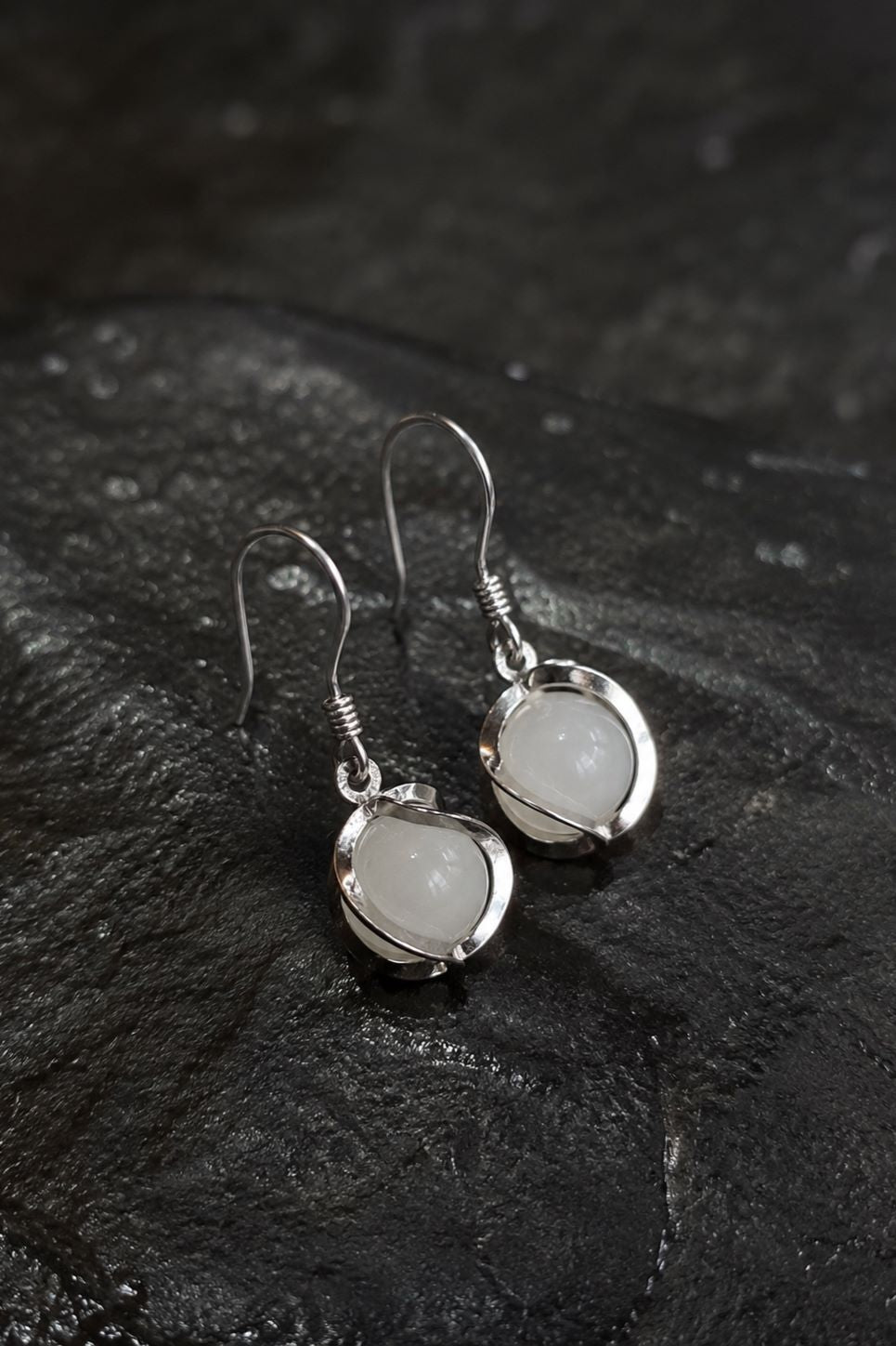 A pair of silver drop earrings with round, milky white stone bezels, displayed on a dark textured slate surface.