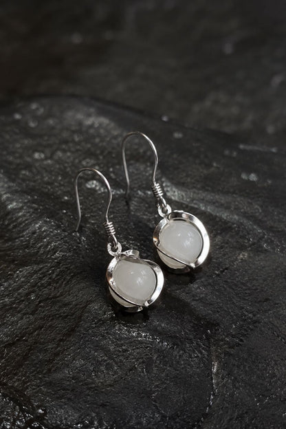 A pair of silver drop earrings with round, milky white stone bezels, displayed on a dark textured slate surface.