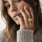 A close-up lifestyle shot of a woman wearing a chunky, textured gold ring on her finger, dressed in a cozy beige knit sweater against a soft, light background.