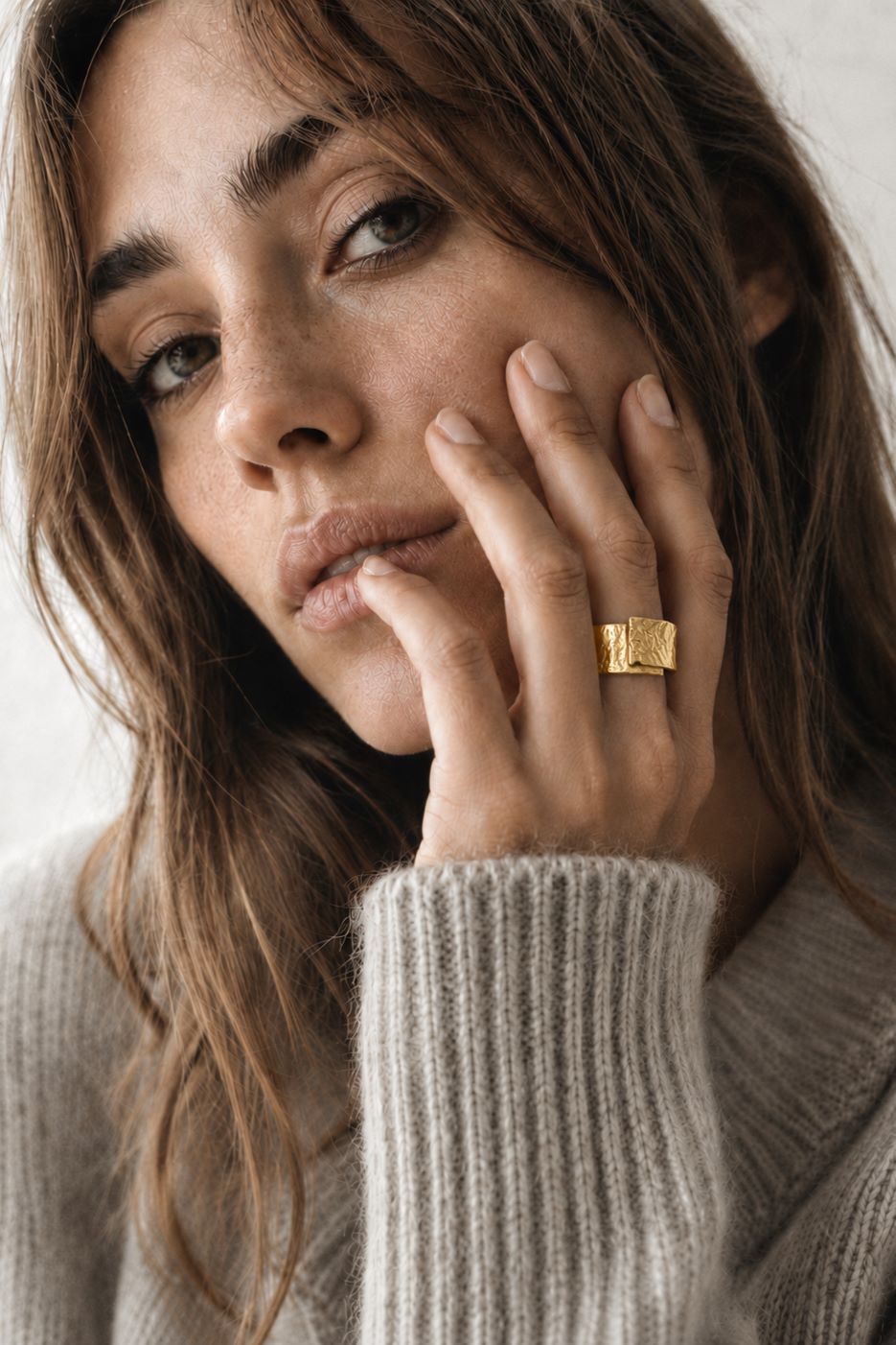 A close-up lifestyle shot of a woman wearing a chunky, textured gold ring on her finger, dressed in a cozy beige knit sweater against a soft, light background.
