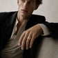 A man wearing a black blazer over a light beige shirt, resting his arm on a textured gray sofa and wearing a wide gold band ring on his finger.