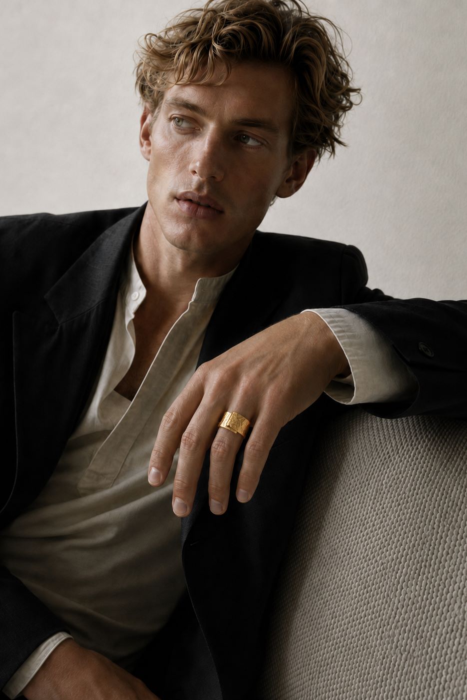 A man wearing a black blazer over a light beige shirt, resting his arm on a textured gray sofa and wearing a wide gold band ring on his finger.