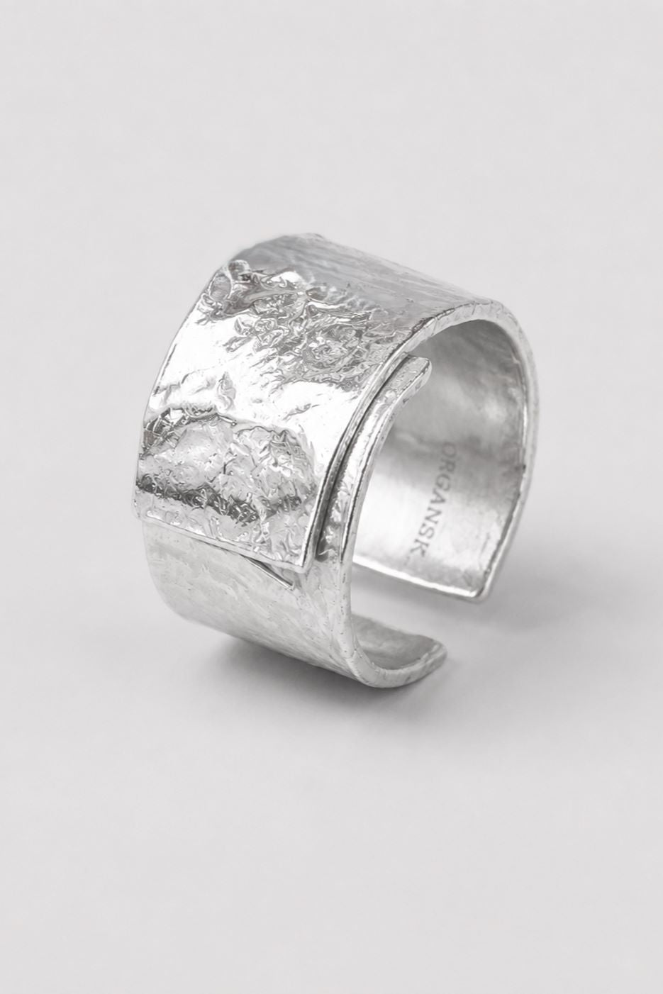 A wide, chunky silver cuff ring with a heavily textured, hammered finish, photographed on a neutral light gray background.