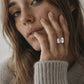 A close-up lifestyle shot of a woman wearing a textured silver ring on her finger, dressed in a cozy beige ribbed knit sweater against a soft, neutral background.