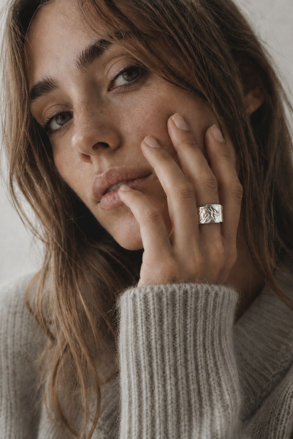 A close-up lifestyle shot of a woman wearing a textured silver ring on her finger, dressed in a cozy beige ribbed knit sweater against a soft, neutral background.