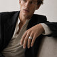 A man wearing a black blazer over a beige shirt, resting his arm on a textured beige sofa and wearing a silver band ring.