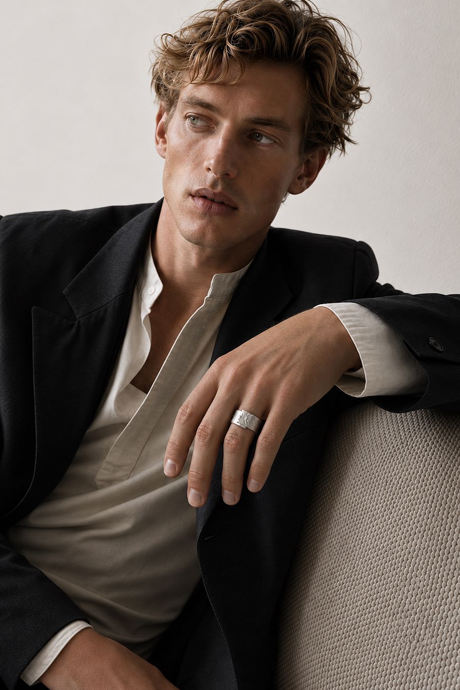 A man wearing a black blazer over a beige shirt, resting his arm on a textured beige sofa and wearing a silver band ring.
