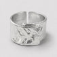 A wide, textured silver ring with a hammered organic finish and an open split design, photographed on a neutral light gray background.