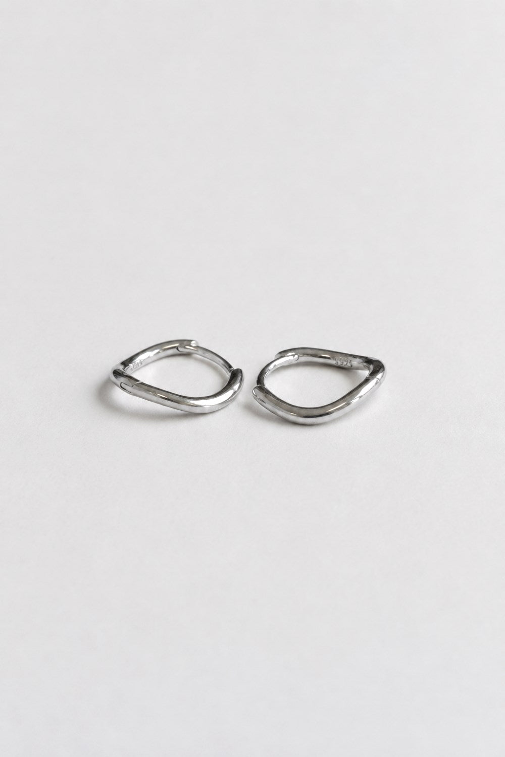 A pair of small, abstract silver hoop earrings with a textured, organic wavy outline, displayed on a clean, light gray background.