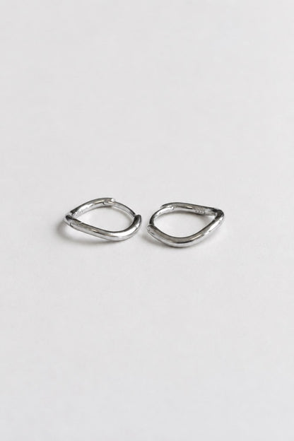 A pair of small, abstract silver hoop earrings with a textured, organic wavy outline, displayed on a clean, light gray background.