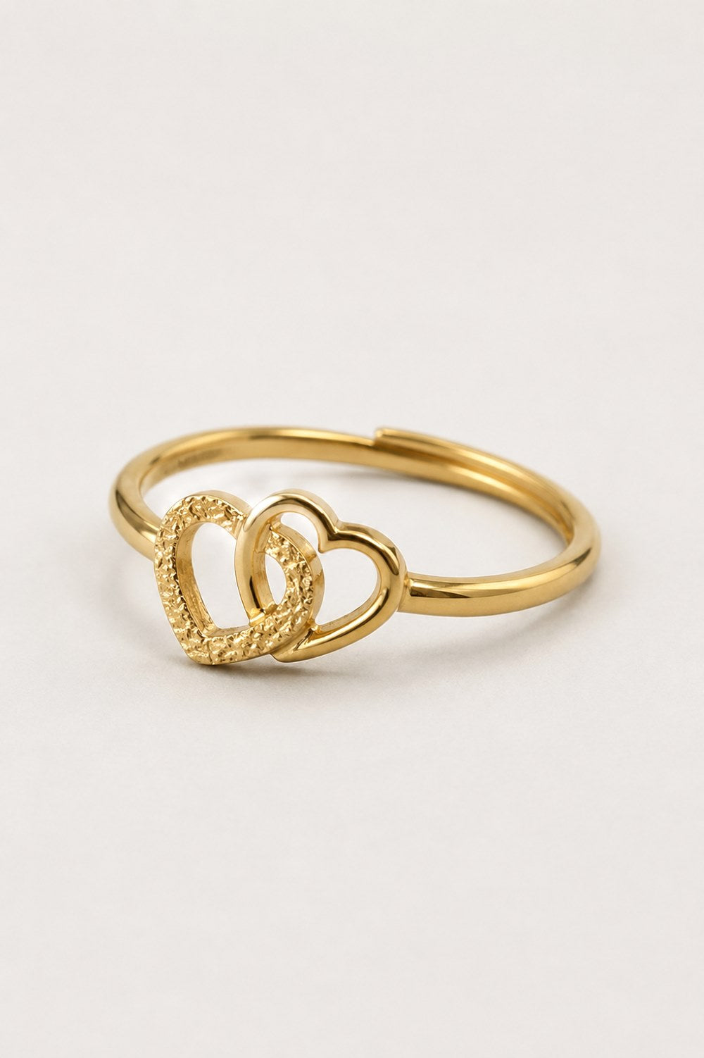 A gold ring featuring two interlocking open heart designs, one with a smooth polished finish and the other with a textured, rope-like border, set against a plain light background.