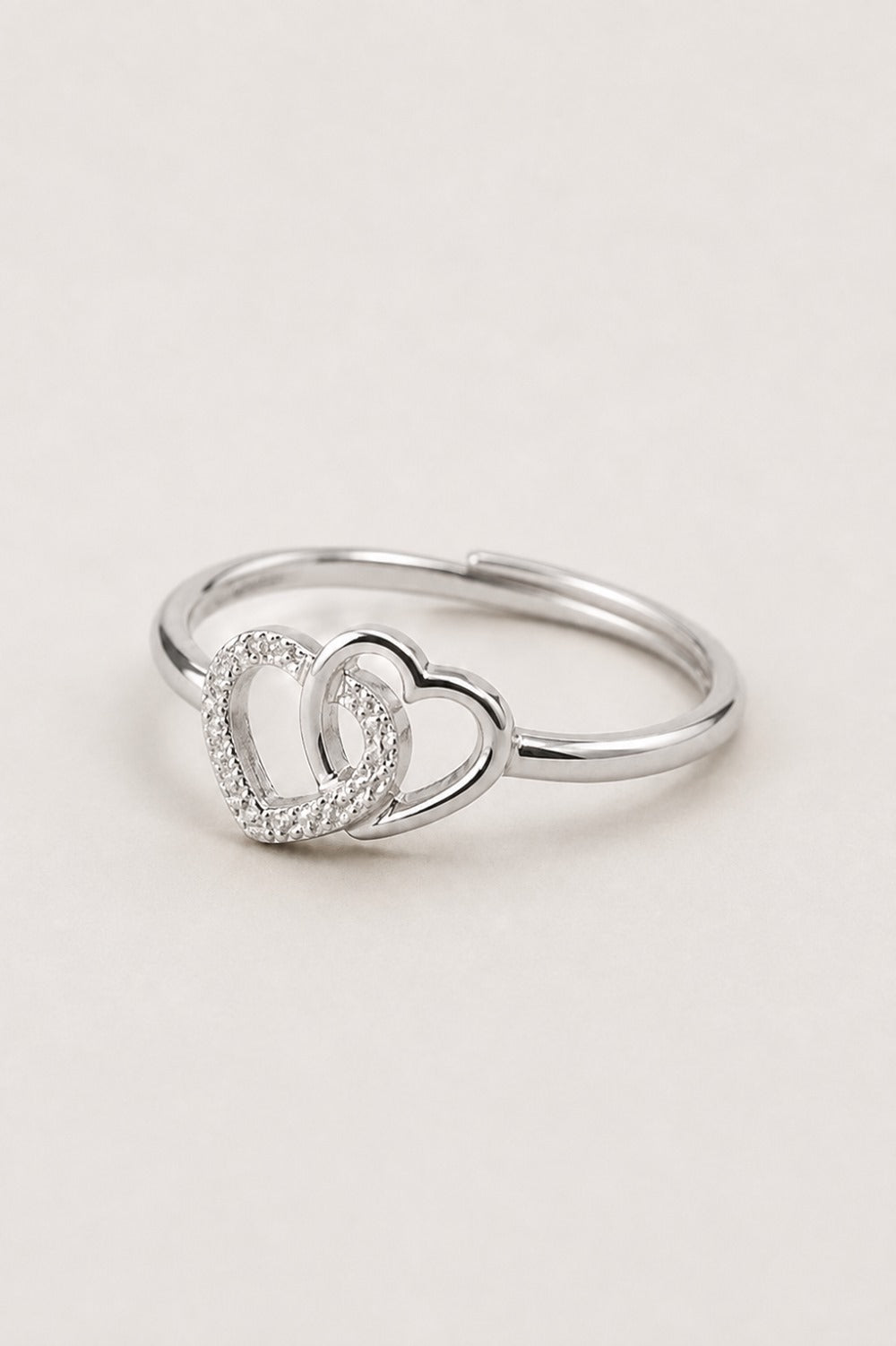 A silver-toned ring featuring two interlocking open heart designs, with one heart encrusted with small clear stones, set against a plain light beige background.