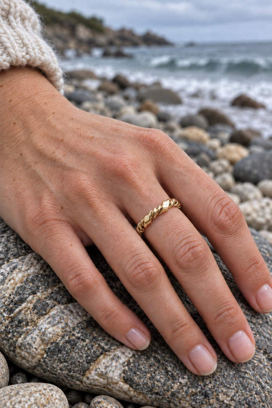 A gold twisted band ring worn on a person's ring finger, resting on a large rock with a rocky beach and ocean waves in the blurred background.