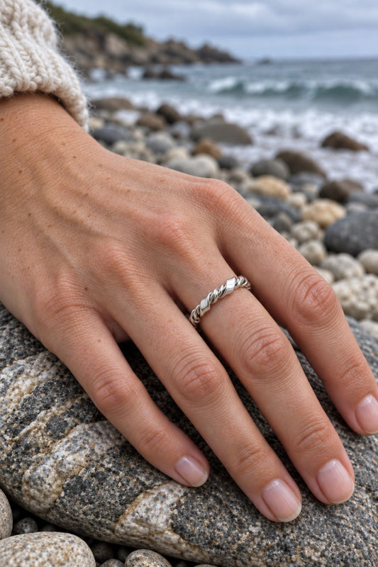 A close-up of a person's hand resting on a rock at the beach, wearing a thin twisted silver band ring.