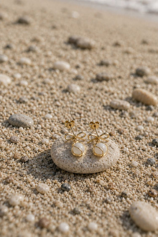 A pair of gold bow and pearl stud earrings resting on a smooth stone on a sandy beach.