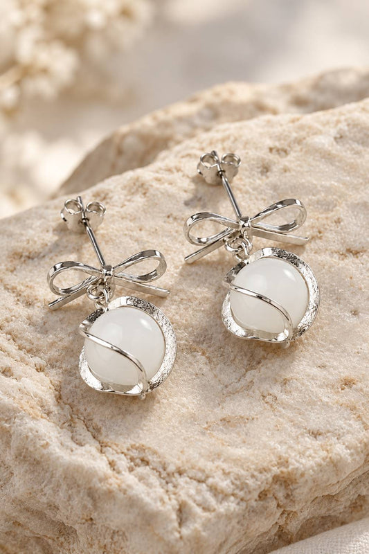 A pair of silver-toned dangle earrings with bow-shaped posts and oval milky white stones, displayed on a textured beige stone surface.