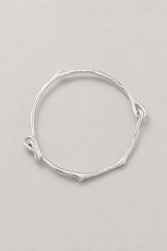 A thin silver bangle bracelet with a textured, twig-like organic shape and small loop details at the ends, displayed on a neutral light gray background.
