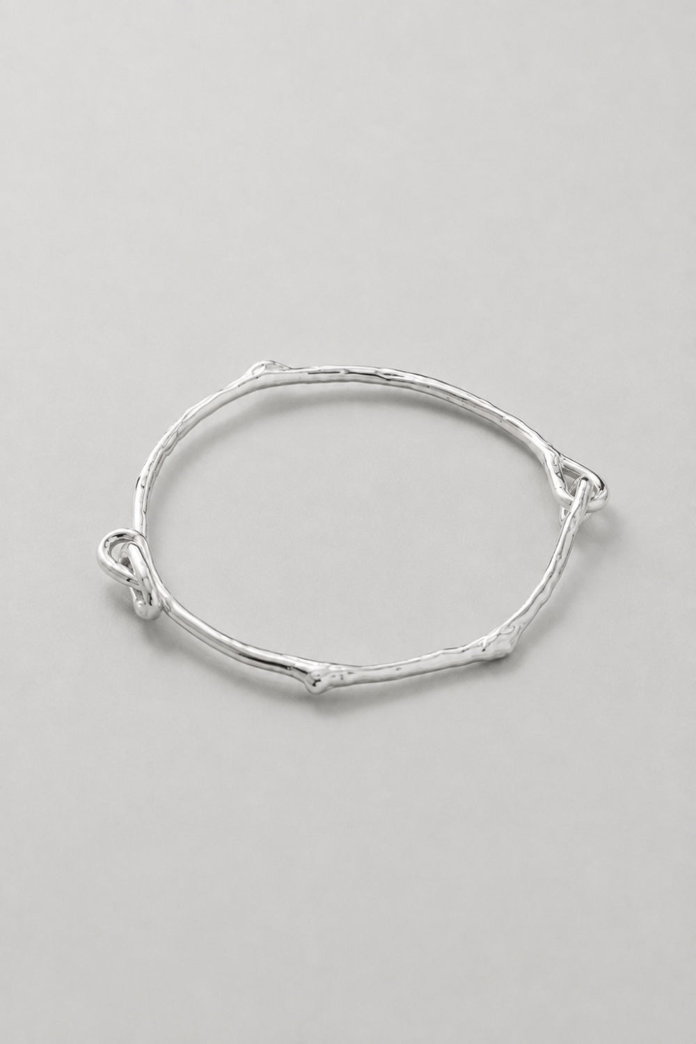 A thin, textured silver bangle bracelet with organic, twig-like branches and a small loop closure, shown on a neutral light gray background.