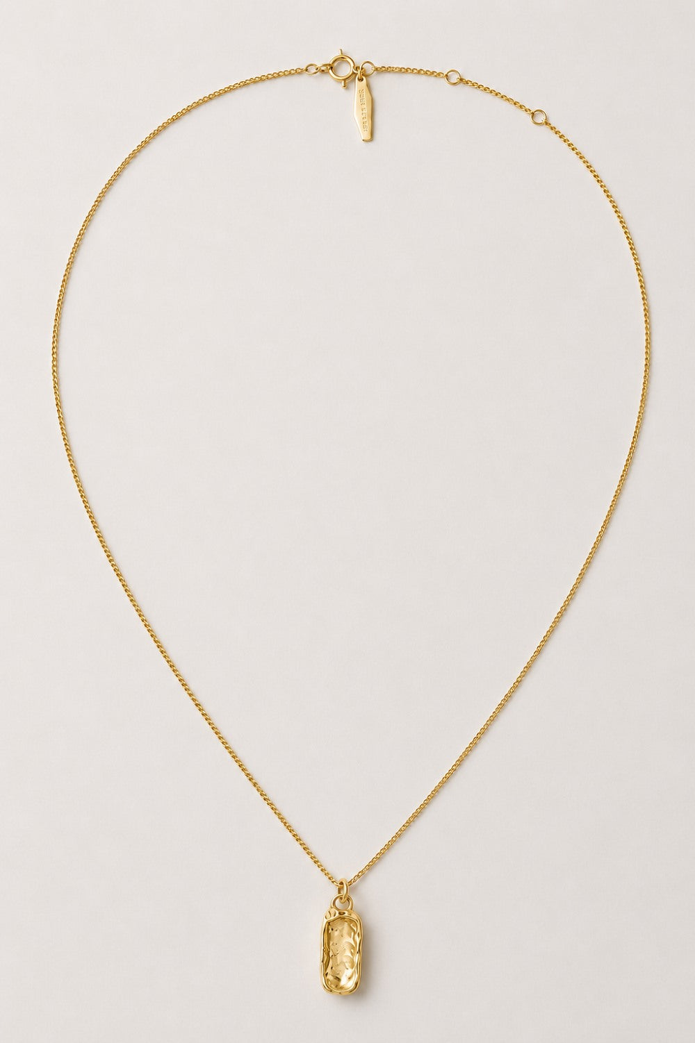 A delicate gold chain necklace with a small, vertically oriented oval-cut gold pendant, displayed on a clean, light gray background.