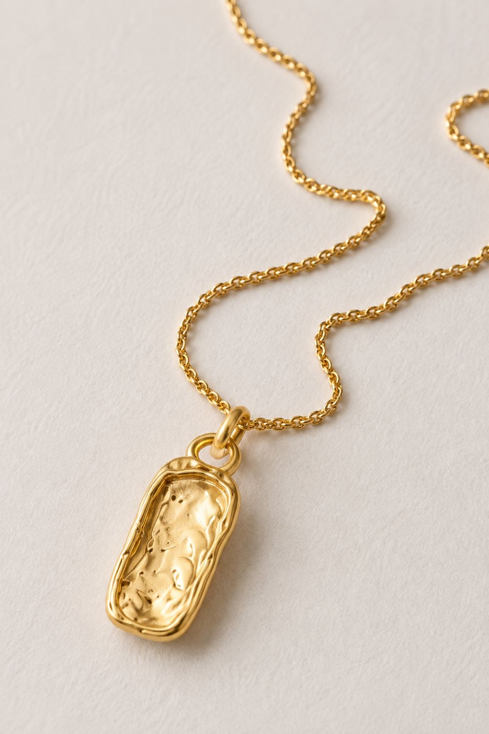 A gold necklace with a textured, irregularly shaped pendant, displayed on a light off-white textured background.