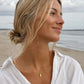 A woman with blonde hair in a low bun wearing a delicate gold pendant necklace, photographed outdoors on a beach with the ocean and cloudy sky in the background.