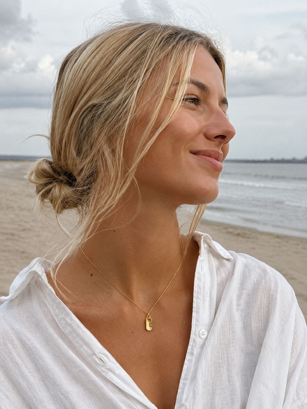 A woman with blonde hair in a low bun wearing a delicate gold pendant necklace, photographed outdoors on a beach with the ocean and cloudy sky in the background.