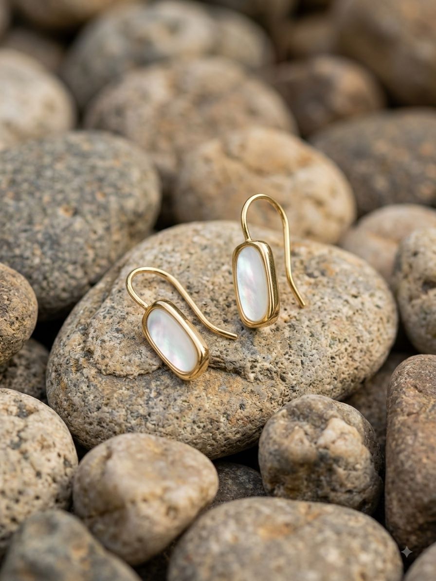 A pair of gold drop earrings with oval white mother-of-pearl inlays, resting on smooth river stones.