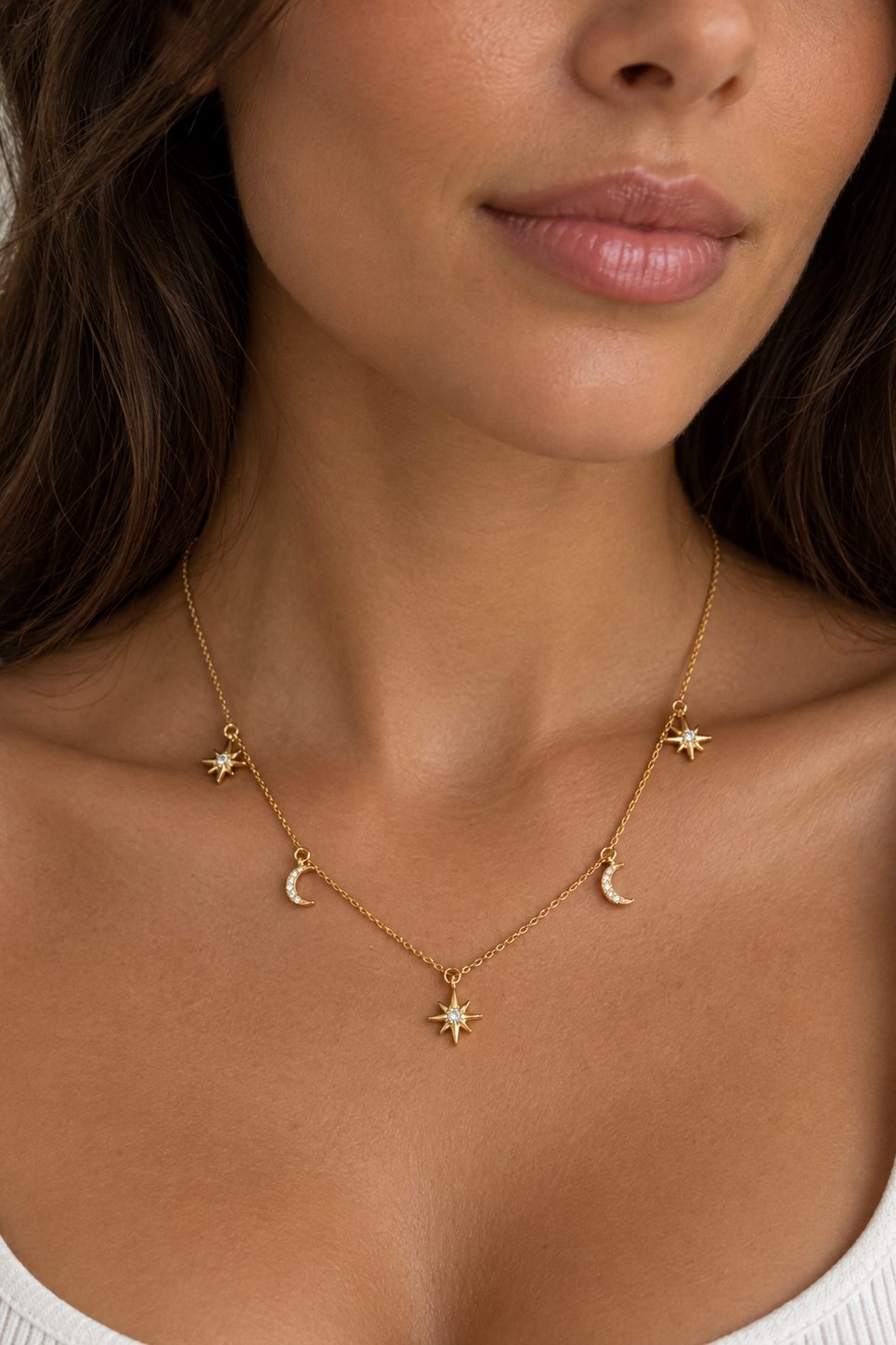 A close-up of a model wearing a delicate gold necklace with small celestial charms, including three crescent moons and three starburst pendants, arranged in a Y-shape.